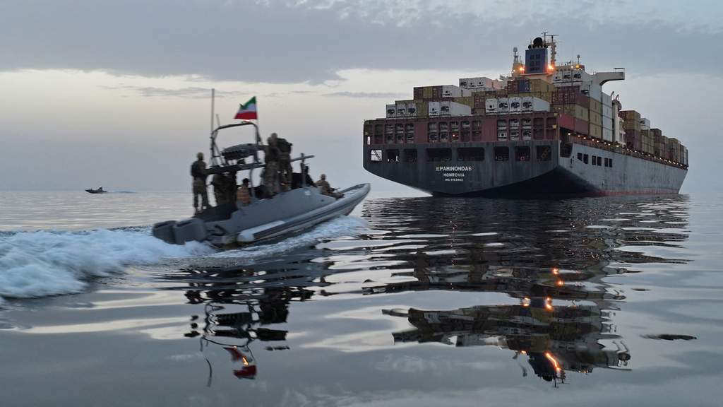 A large container cargo ship navigating through a narrow maritime chokepoint under high-security watch and rough open seas.