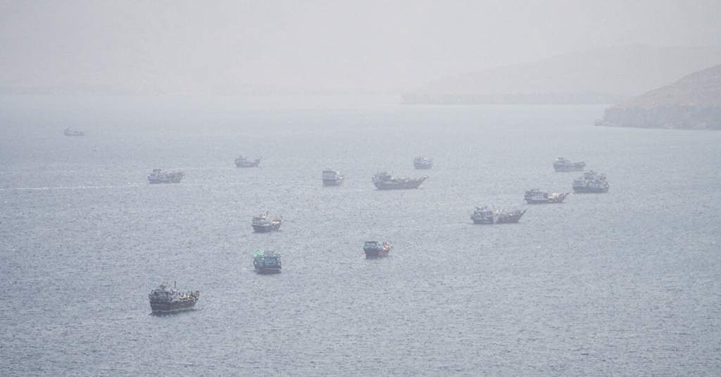 A cargo ship navigating rough waters in the Middle East with smoke rising in the background near a strategic maritime oil shipping chokepoint.